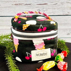 May include: A black fabric cosmetic bag with a zipper closure and a top handle. The bag features a repeating pattern of pink ice cream cones and red cherries. A rectangular white label on the front reads Sigi Stardust Handmade Happiness.