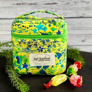 May include: A yellow fabric lunch bag featuring a vibrant blue and green floral pattern. The bag has a top handle, a green zipper closure, and a rectangular white label on the front with the text Sigi Stardust Handmade Happiness.