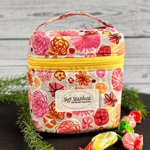 May include: A small, structured fabric bag with a top handle and a yellow zipper. The bag features a floral pattern in shades of pink, orange, and yellow on a light gray background. A rectangular label on the front reads Sigi Stardust Handmade Happiness.