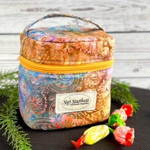May include: A small, square fabric storage bag with a handle, featuring a colorful batik-style pattern in shades of blue, orange, and pink. It has a bright yellow zipper and a white rectangular label reading Sigi Stardust Handmade Happiness.