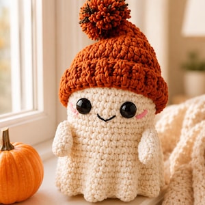 May include: A crocheted ghost figurine featuring a cream-coloured body and a textured burnt orange beanie topped with a matching pom-pom. The ghost has large black plastic eyes, a small stitched smile, and subtle pink embroidered cheeks.