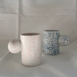 May include: Two ceramic mugs with speckled glaze. One mug is pink and the other is blue. Both mugs have a rounded handle.