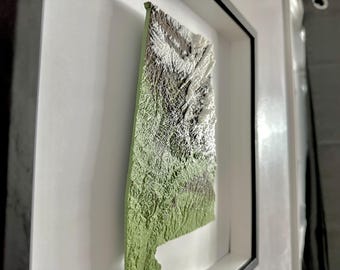 Può includere: Una mappa in rilievo 3D incorniciata di uno stato, che mostra dettagli topografici con sfumature di verde, grigio e bianco. La mappa è montata all'interno di una cornice bianca con un bordo nero, creando un effetto a strati. L'opera d'arte è esposta ad angolo.
