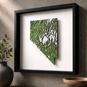 May include: A framed three-dimensional wall art piece depicting the state of Nevada. The map features a textured green surface with raised white topographical details representing mountain ranges, all encased in a simple black square frame.