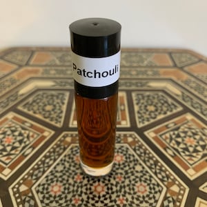 May include: A clear glass roll-on bottle filled with a dark amber liquid. The bottle has a black cap and a white label with the word "Patchouli" printed on it. The bottle is displayed on a decorative table.