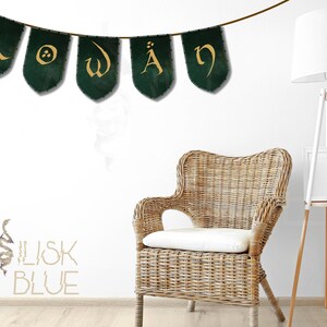 May include: A green and gold banner with the word "ROWAN" written in a decorative font. The banner is hanging on a white wall with a wicker chair and a white floor lamp in the background. The words "BASILISK BLUE" are written in a decorative font on the wall below the banner.