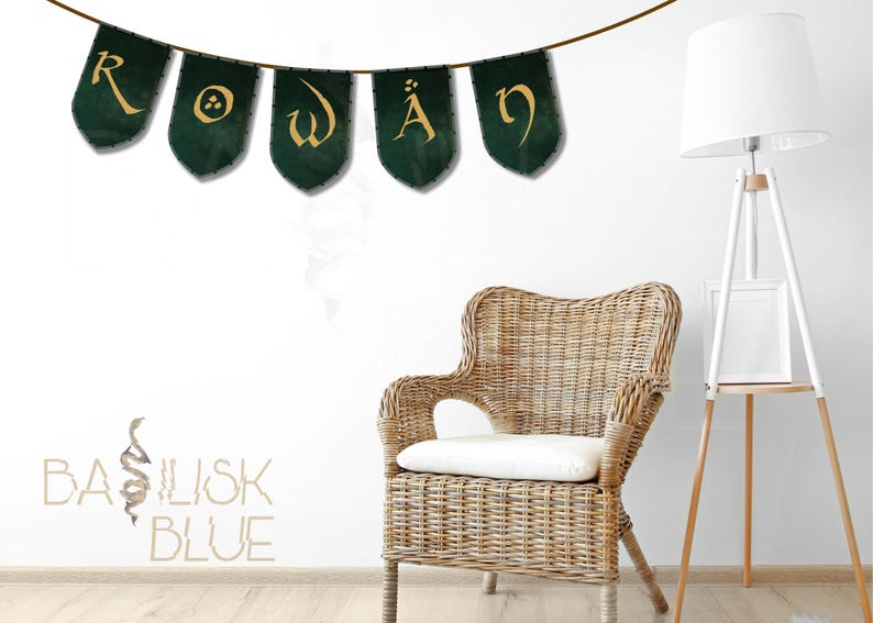 May include: A green and gold banner with the word "ROWAN" written in a decorative font. The banner is hanging on a white wall with a wicker chair and a white floor lamp in the background. The words "BASILISK BLUE" are written in a decorative font on the wall below the banner.