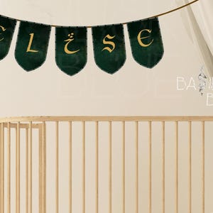 May include: A green and gold banner with the letters "ELISE" in a decorative font. The banner is hanging above a wooden crib.