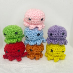 May include: Six crocheted octopus plushies in different colors, including pink, green, blue, purple, red, and yellow. The plushies are stacked on top of each other.