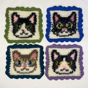 May include: Four square crochet coasters, each featuring a cat face design. The coasters have scalloped edges in green, blue, navy, and purple. The cat faces are black, brown, and white with pink noses and ears, and green or blue eyes.
