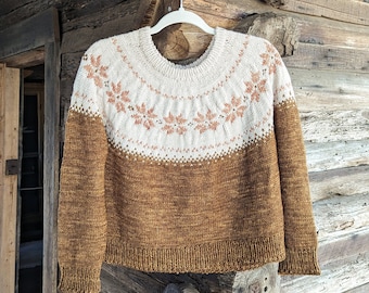 May include: A brown and white knitted sweater with a geometric pattern. The sweater is hanging on a wooden hanger against a rustic wooden background.