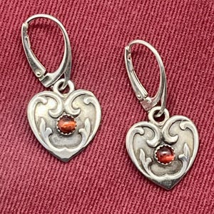 May include: A pair of silver heart-shaped earrings with red gemstone accents. The earrings have a delicate filigree design and dangle from a hook.