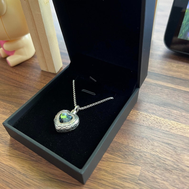 May include: A silver heart-shaped pendant necklace with a green gemstone in the center. The necklace is in a black velvet box.