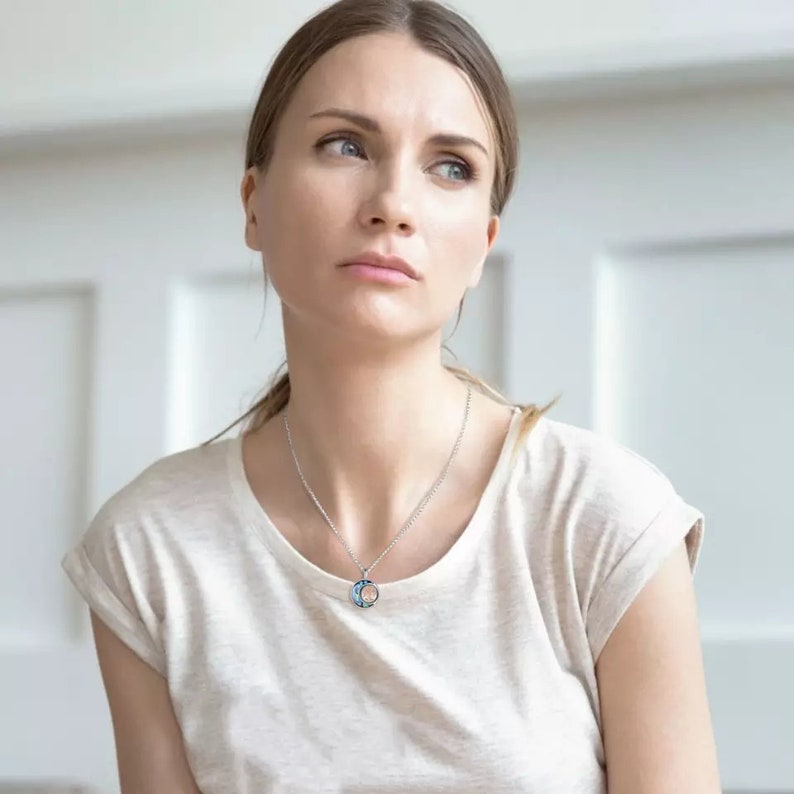 Pu&ograve; includere: Una donna indossa una maglietta bianca con una collana d'argento con un ciondolo blu e argento. Il ciondolo &egrave; un cerchio con un disegno all'interno.