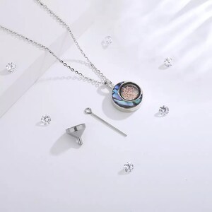 Pu&ograve; includere: Una collana d'argento con un ciondolo rotondo che presenta un design ad albero della vita. Il ciondolo &egrave; realizzato in madreperla di abalone con un design ad albero rosa. La collana viene fornita con un piccolo imbuto e uno spillo d'argento.