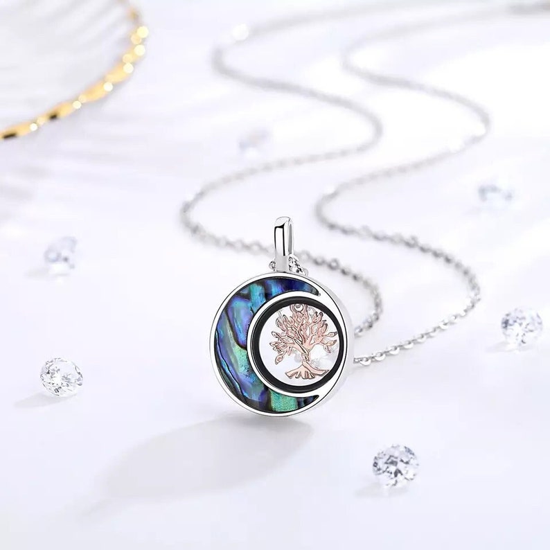 Pu&ograve; includere: Una collana d'argento con un ciondolo a forma di luna crescente e un albero della vita. Il ciondolo &egrave; realizzato in madreperla di abalone con una finitura blu, verde e iridescente. L'albero della vita &egrave; in oro rosa.