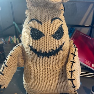 May include: A knitted brown and black Oogie Boogie doll with a stitched smile and eyes. The doll is standing on a wooden base.