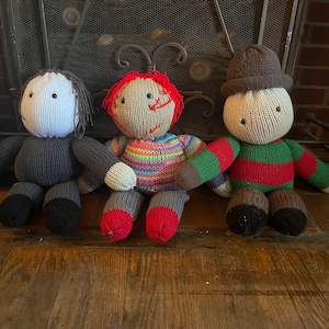 Puede incluir: Tres muñecas de punto, cada una con un diseño diferente. La muñeca de la izquierda es gris con la cara blanca y los ojos negros. La muñeca del centro lleva una camisa a rayas de arcoíris y tiene la cara roja con rasgos cosidos. La muñeca de la derecha lleva una camisa a rayas verdes y rojas y un sombrero marrón.