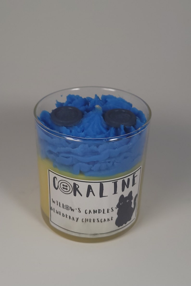 May include: A blue and yellow candle in a glass jar with a black cat silhouette and the text "CORALINE WILLOW'S CANDLES BLUEBERRY CHEESECAKE".