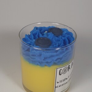 May include: A glass jar candle with a yellow and blue layered wax design. The top layer is blue with two blue wax blueberries. The label reads "CORAL & WILLOWS CANDLE CO. BLUEBERRY LEMONADE".