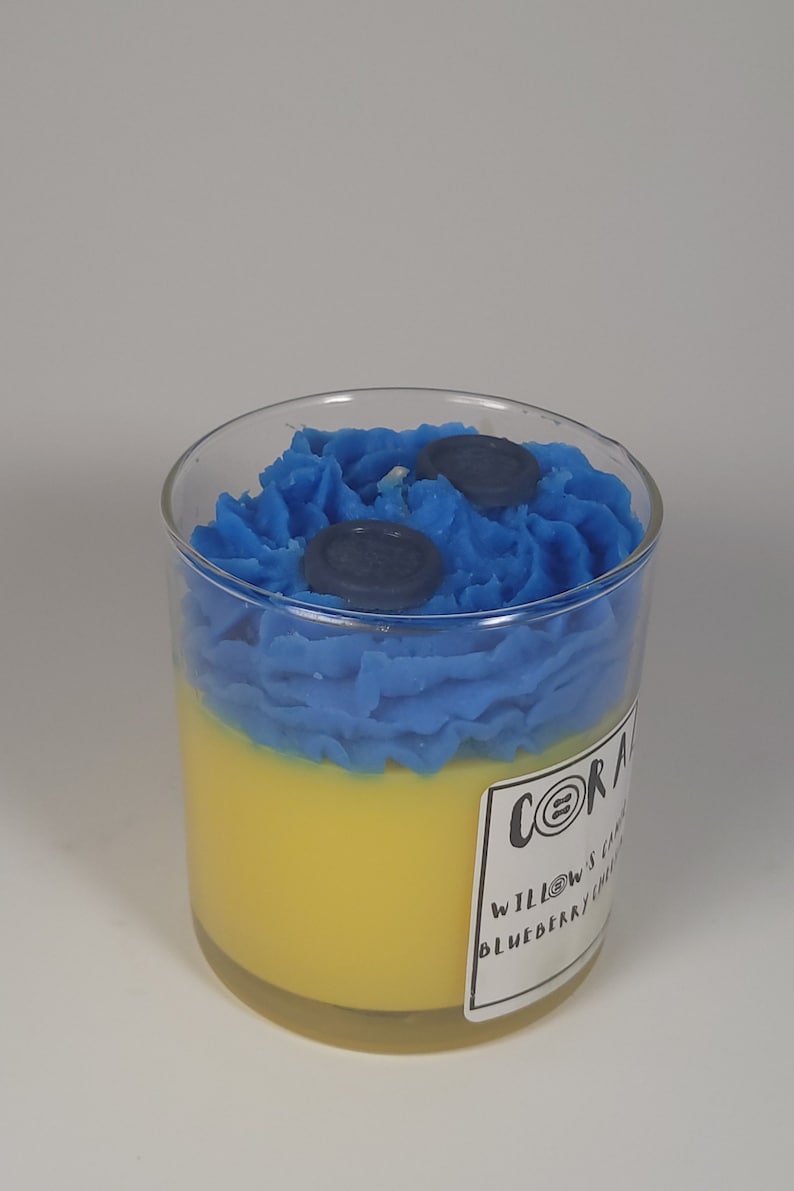 May include: A glass jar candle with a yellow and blue layered wax design. The top layer is blue with two blue wax blueberries. The label reads "CORAL & WILLOWS CANDLE CO. BLUEBERRY LEMONADE".