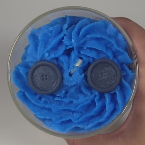 May include: A blue candle with two dark blue buttons on top. The candle is in a clear glass jar.