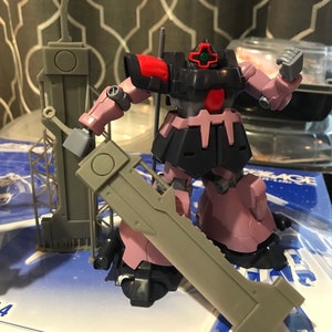 May include: A gray and pink robot action figure with a large gray sword. The robot has a red and black head with a green cross on the forehead. The robot is standing on a blue and white box with the text "Gundam Age-2 Arthur" in Japanese and English.