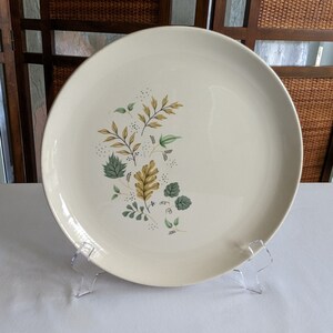 May include: A white ceramic dinner plate with a floral design featuring green and yellow leaves. The plate has a slightly raised rim and is decorated with a stylized floral pattern.