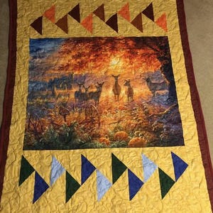 May include: A decorative quilt with a central image of deer in a forest at sunset. The quilt has a yellow background with triangle patterns in orange, brown, blue, and green. The border is a deep red.
