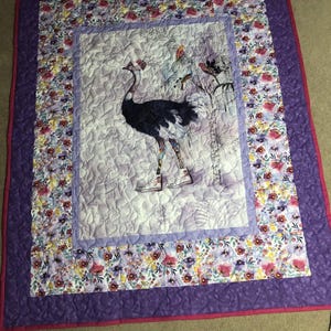 May include: A quilted blanket with a central image of a stylized ostrich wearing boots and a hat. The quilt features a floral border in shades of purple, pink, and white, with a solid purple backing and a pink trim. The ostrich is depicted in black and white.