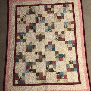 Pode incluir: Uma colcha de retalhos com fundo branco e uma variedade de padrões florais em tons de marrom, rosa e azul. A colcha é enfeitada com uma borda rosa e bordô.