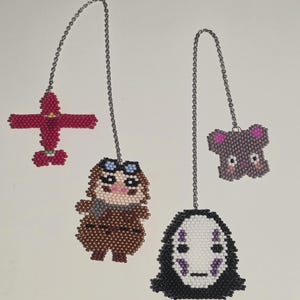 May include: Four beaded charms hang from silver chains. A red airplane, a brown figure with goggles, a purple koala, and a black and white face are the subjects. The charms are made of small, colorful beads.