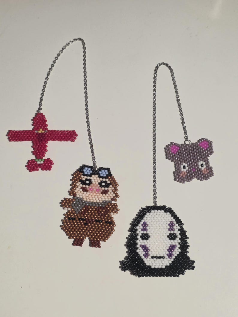 May include: Four beaded charms hang from silver chains. A red airplane, a brown figure with goggles, a purple koala, and a black and white face are the subjects. The charms are made of small, colorful beads.