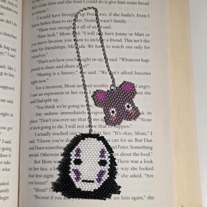 May include: A handmade beaded bookmark featuring two designs: a No-Face character and a bear. The No-Face design has a white face with black and purple details, while the bear is purple with pink accents. The bookmark is attached to a silver chain.