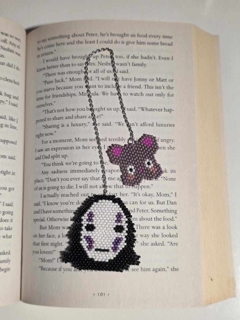 May include: A handmade beaded bookmark featuring two designs: a No-Face character and a bear. The No-Face design has a white face with black and purple details, while the bear is purple with pink accents. The bookmark is attached to a silver chain.