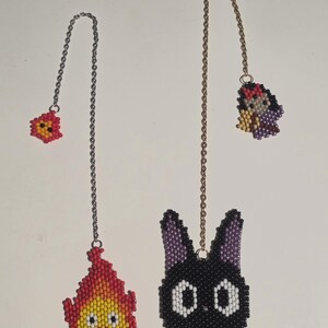May include: Two beaded bookmarks featuring cartoon characters. One is a red and yellow flame, the other a black cat with white eyes and a red bow. Both have chain attachments.