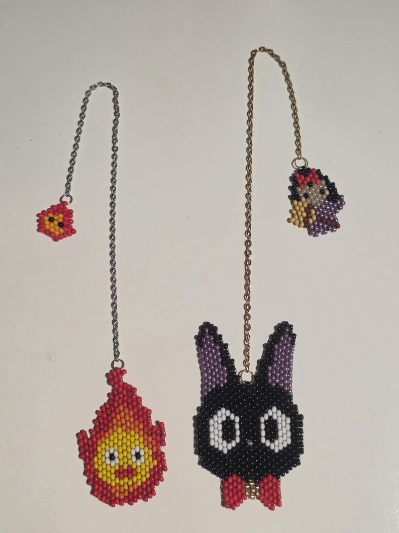 May include: Two beaded bookmarks featuring cartoon characters. One is a red and yellow flame, the other a black cat with white eyes and a red bow. Both have chain attachments.