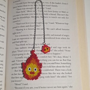 May include: A handmade beaded bookmark featuring a smiling flame design in red, yellow, and orange. The bookmark is attached to a silver chain and is resting on the pages of a book.