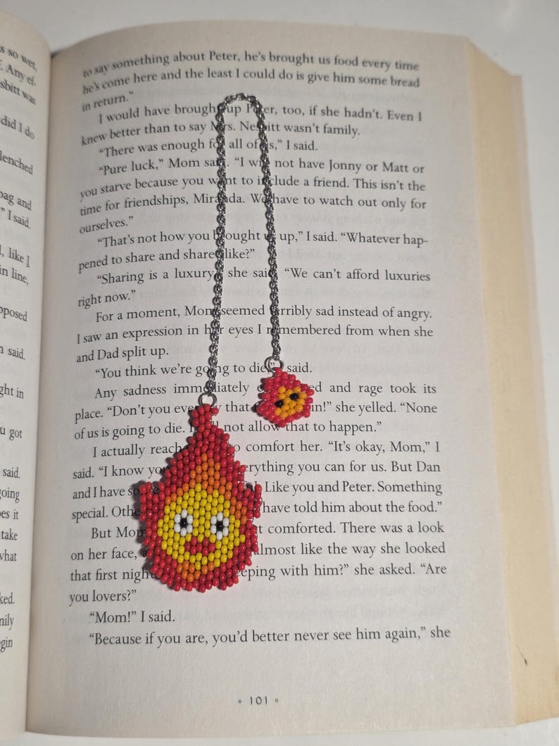 May include: A handmade beaded bookmark featuring a smiling flame design in red, yellow, and orange. The bookmark is attached to a silver chain and is resting on the pages of a book.