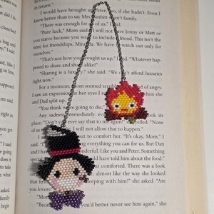 May include: Two beaded bookmarks with chain attachments. One features a character with a black top hat, a white face, and a purple bow tie. The other is a red, yellow, and orange flame design. Both are on a book page.