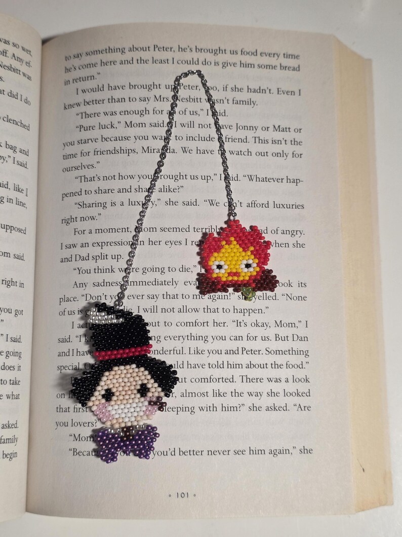 May include: Two beaded bookmarks with chain attachments. One features a character with a black top hat, a white face, and a purple bow tie. The other is a red, yellow, and orange flame design. Both are on a book page.