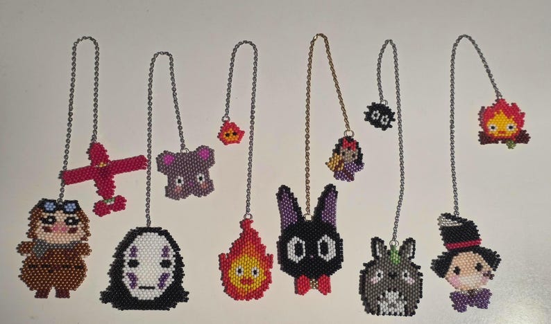 May include: A collection of beaded anime-themed charms, each suspended by a silver chain. The charms feature characters like No-Face, Totoro, and Calcifer, rendered in vibrant colors and pixelated designs. The collection includes a red airplane and a purple koala.