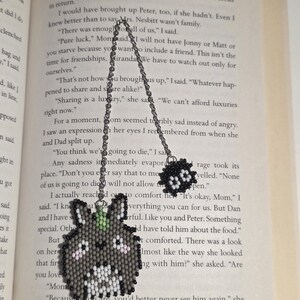 May include: A handmade beaded Totoro-inspired pendant necklace. The pendant features a gray Totoro design with white, black, and pink accents, and a small green leaf. The necklace has a silver chain and a small black beaded charm.