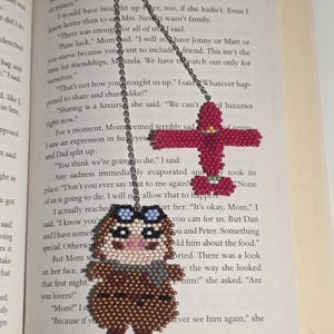 May include: Handmade beaded owl bookmark with aviator goggles and a scarf, and a red beaded cross bookmark. The owl is brown, tan, and gray. The bookmarks are hanging on a book page.