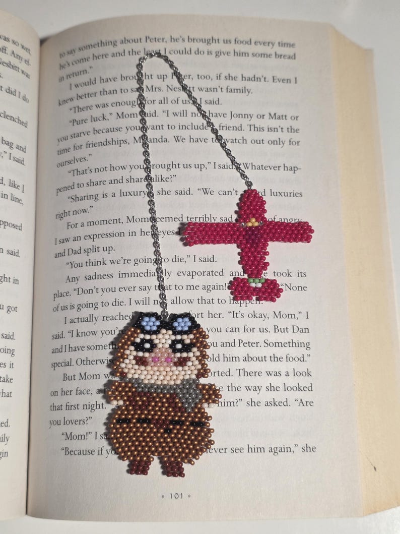 May include: Handmade beaded owl bookmark with aviator goggles and a scarf, and a red beaded cross bookmark. The owl is brown, tan, and gray. The bookmarks are hanging on a book page.