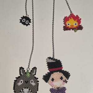 May include: Four beaded charms hang from silver chains. The charms include a black soot sprite, a red and yellow fire demon, a gray bunny-like creature, and a character in a top hat and purple bow tie. The charms are likely for jewelry or keychains.