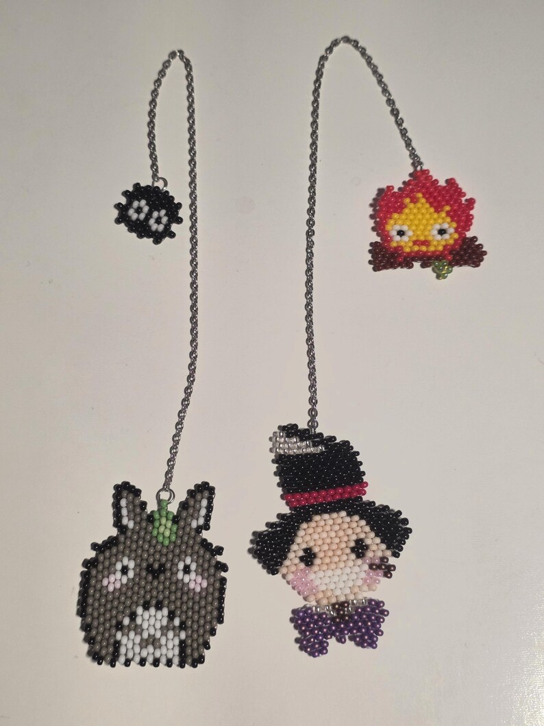 May include: Four beaded charms hang from silver chains. The charms include a black soot sprite, a red and yellow fire demon, a gray bunny-like creature, and a character in a top hat and purple bow tie. The charms are likely for jewelry or keychains.