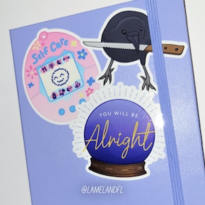May include: Three stickers on a purple background. The first sticker is pink and features a cartoon Tamagotchi with the text "Self Care". The second sticker is a black crow holding a knife. The third sticker is a purple and blue crystal ball with the text "You Will Be Alright" in gold lettering.