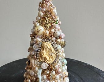 May include: A decorative Christmas tree made of various pearls, beads, and embellishments in shades of white, cream, gold, and pink. The tree features a central cameo and is topped with a decorative floral element. The tree is approximately 6 inches tall.