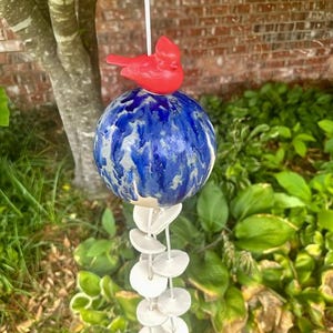 May include: A ceramic wind chime featuring a red cardinal perched atop a blue and white globe. Below the globe are several white, circular chimes. The wind chime hangs from a white string, suitable for outdoor decor.